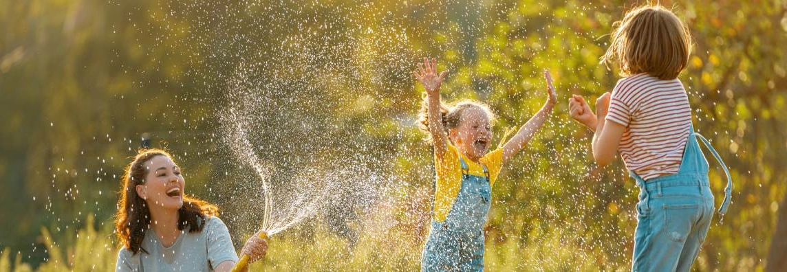 Ein Mensch spritzt zwei Kinder mit einem Gartenschlauch nass.